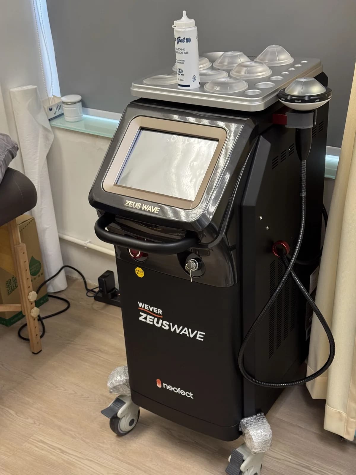 Shockwave unit labelled WEVER ZEUS WAVE on a wheeled cart in a treatment room, with one applicator in its holder and translucent caps and gel on the top tray